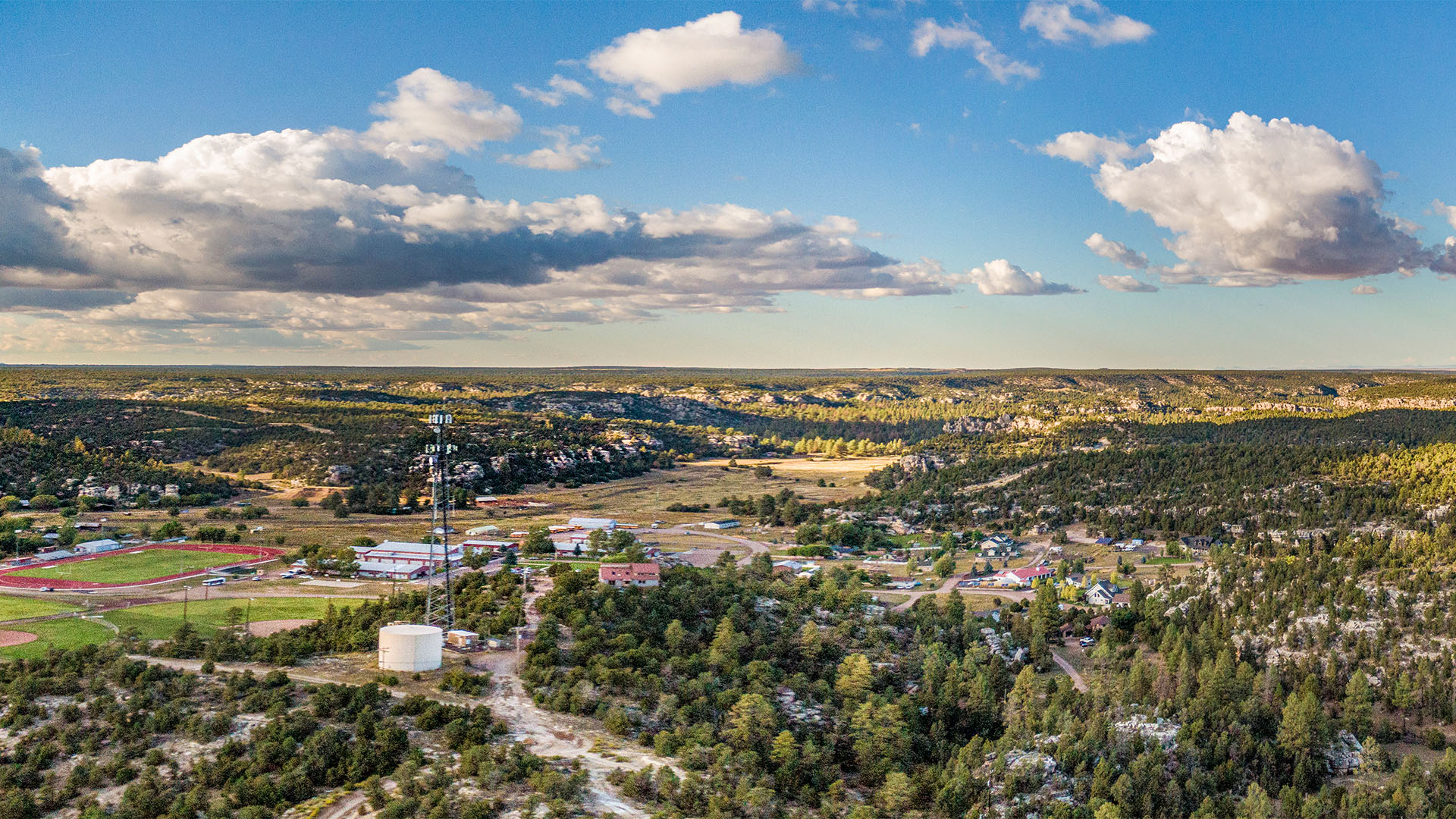 Overlook view of Heber-Overgaard, Arizona, with scattered buildings, a school track, forested hills, and open terrain beneath a partly cloudy sky.